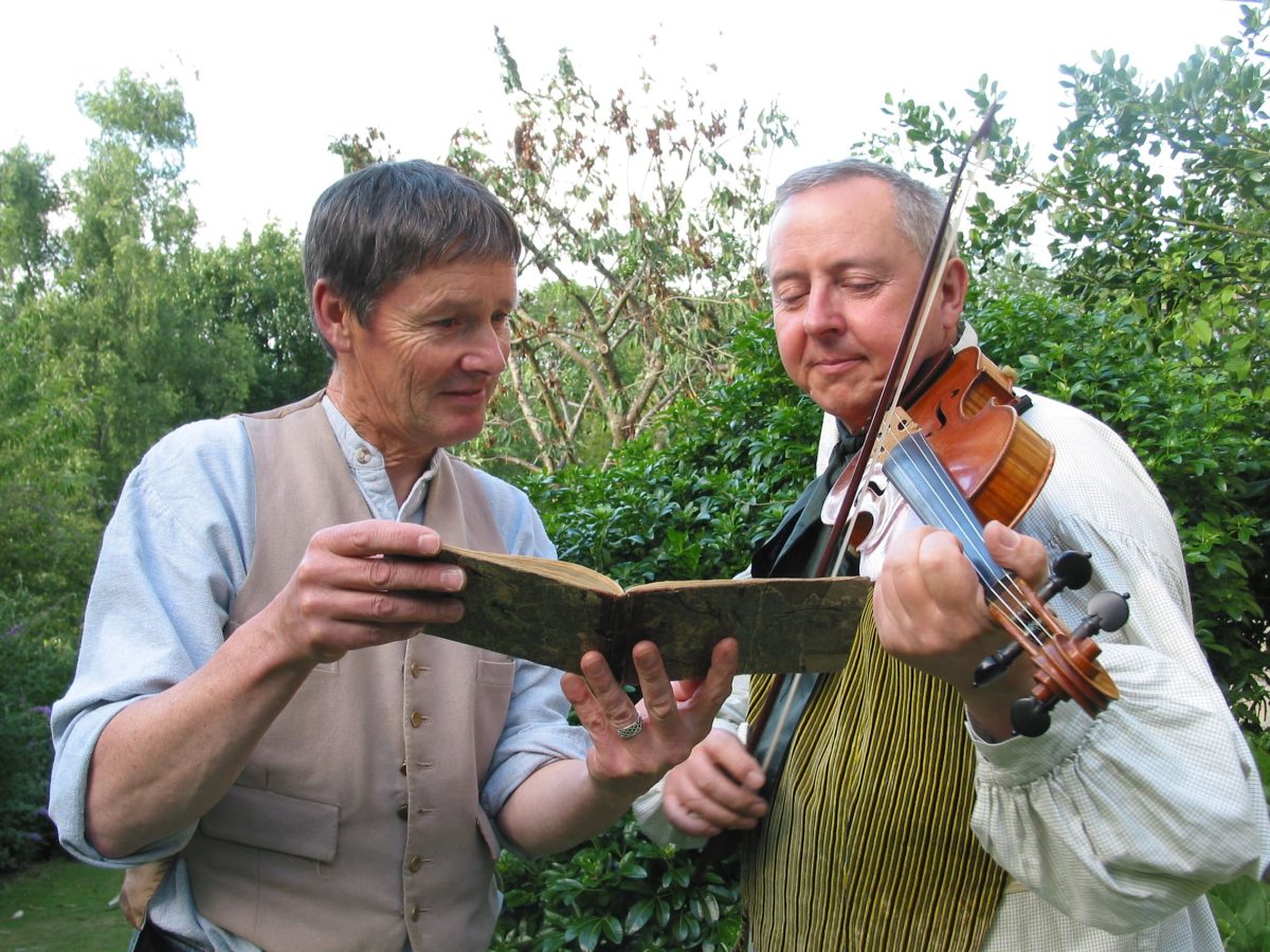 Two folk musicians, one with fiddle, look at open book