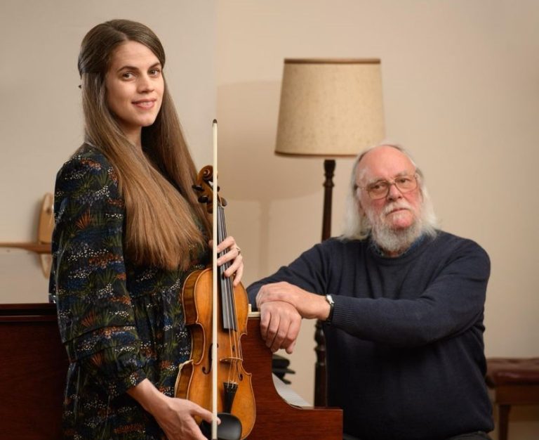 Woman holding violin with a man sat at the piano
